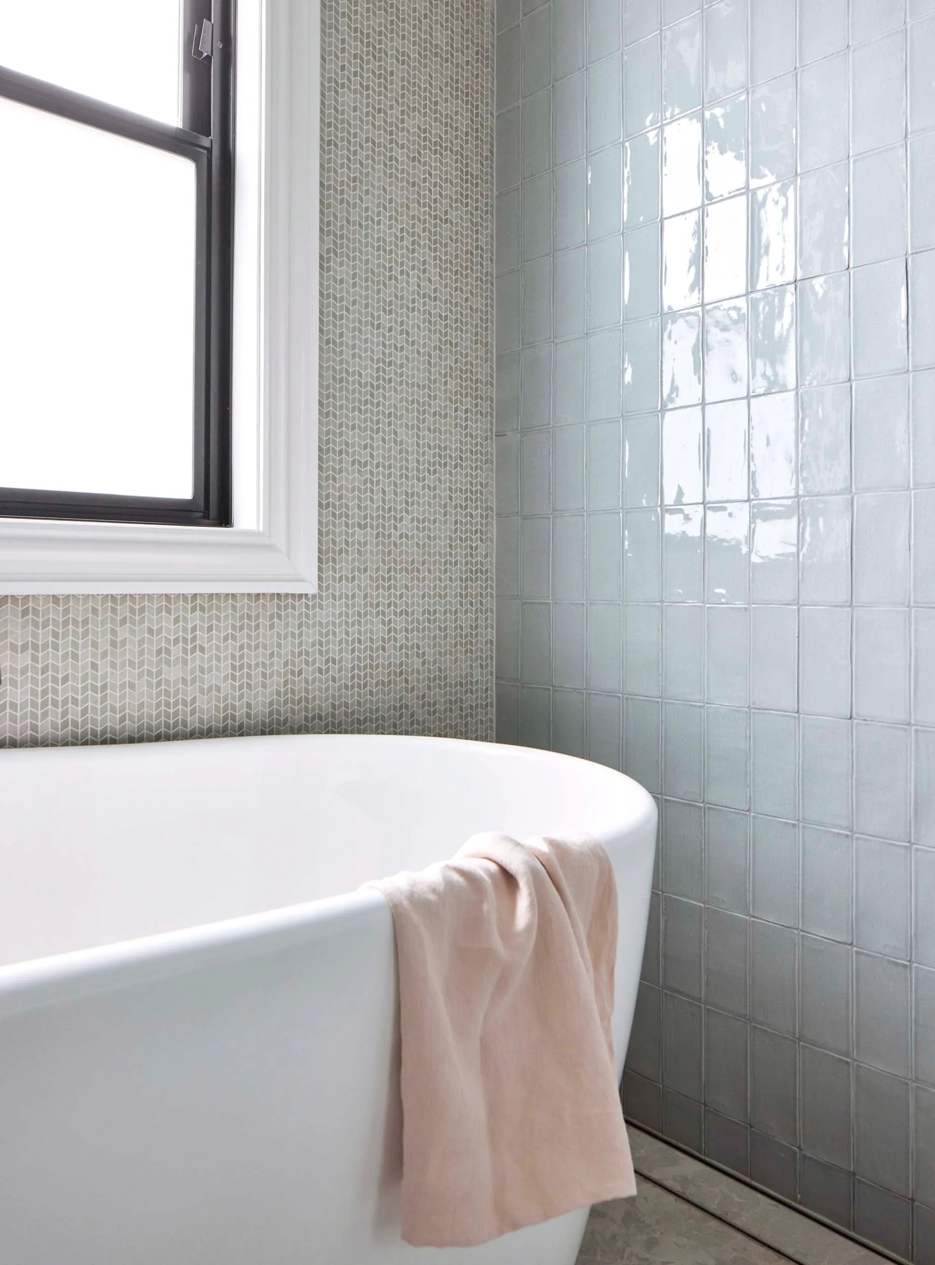 A white bath witha pink handtowel draped over the side sits beneath a window and beside a feature wall of pastel blue tiles