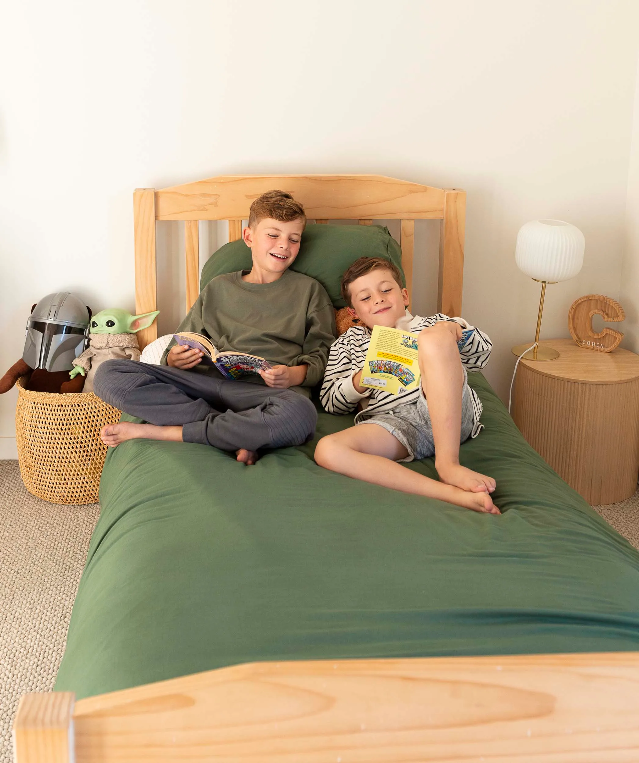 Kids sitting on the single bed with green bedding, a wooden bedframe and a wooden bedside table