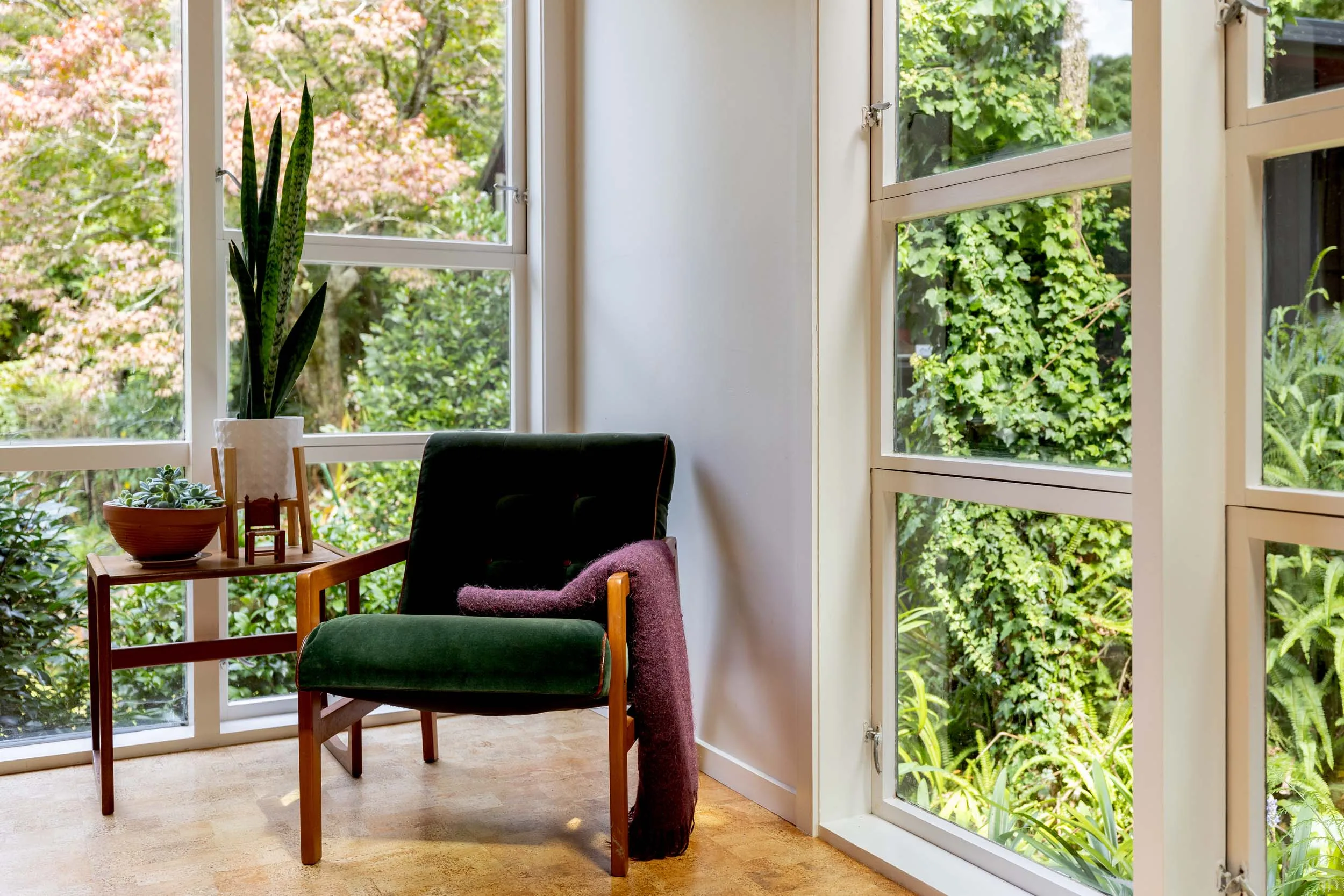 A green fabric chair in the corner of the dining room, with floor-to-ceiling windows on each wall