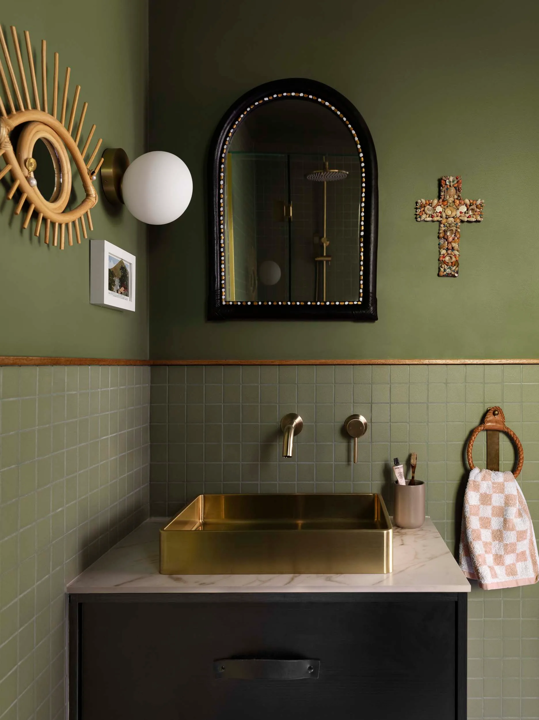 An olive green bathroom vanity with a gold sink and arched mirror
