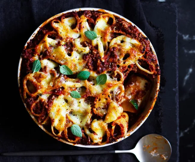 Baked pasta with melted cheese, meat sauce, and fresh basil in a round baking dish, placed on a dark surface.
