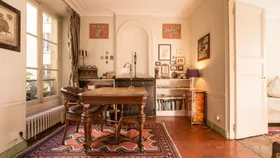 A cozy study room with a wooden table, chairs, and bookshelves, decorated with framed art and a colorful rug.