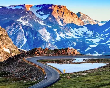 Mountain road with a sharp curve, surrounded by rocky peaks and patches of snow, under a clear blue sky.