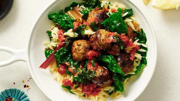 A white bowl of orzo pasta with meatballs, sautéed greens, and tomato sauce.