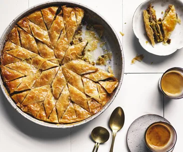 A round metal tin with baklava, cut into an intricate pattern of diamond slices, surrounded by cups of coffee, on a white background.