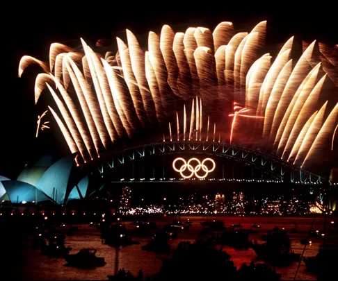 The Sydney Harbour Bridge at nighttime, with a neon-light of the Olympic Games logo, and yellow fireworks firing from the top of the bridge.