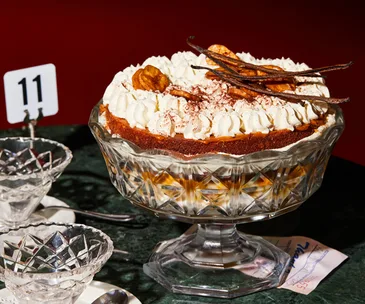 Ornamental glass bowl on a stand holding a trifle topped with whipped cream, banana chips and vanilla bean pods