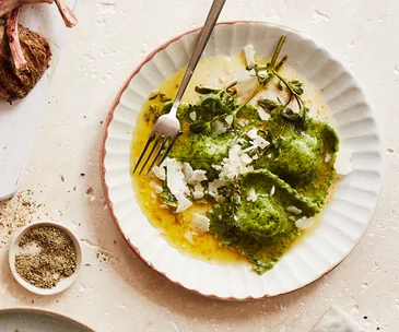 Round, white, ridged plate holding three squares of green ravioli with a butter sauce, and cheese crumbled over the top. 
