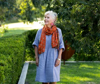 Maggie Beer in a blue smock dress and orange scarf, standing in a garden, looking up and away from the camera