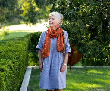 Maggie Beer in a blue smock dress and orange scarf, standing in a garden, looking up and away from the camera