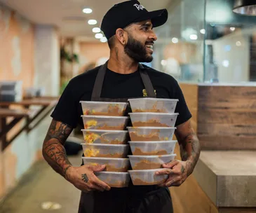 A man in a black T-shirt and black cap, holding two stacks of plastic containers filled with cooked meals.