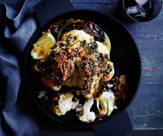 Roasted cauliflower with herbs and spices in a black pan, garnished with lemon wedges on a rustic surface.