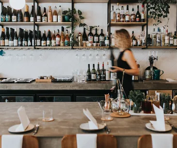 Blonde woman holding a bottle of wine walking behind a restaurant counter.