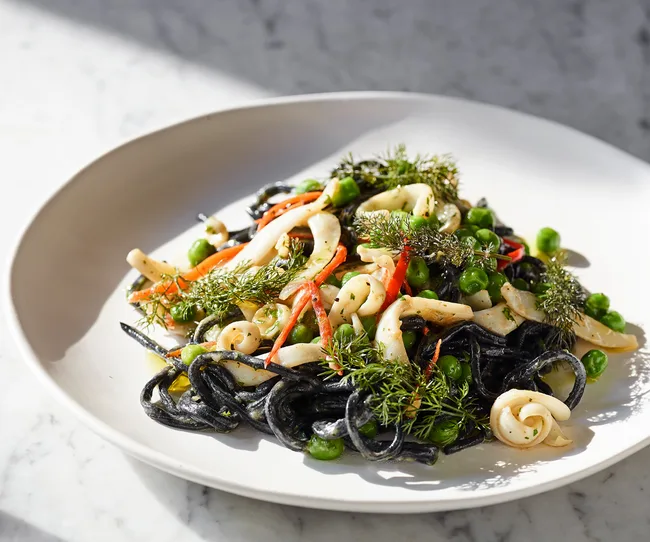 A plate of squid ink pasta with squid, peas, and herbs on a white plate.