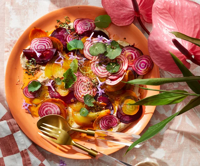 A vibrant salad with sliced beets on an orange plate, decorated with greens and purple flowers, set on a patterned tablecloth.