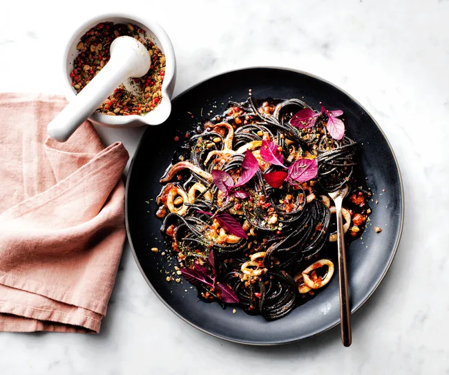 Squid ink pasta with calamari and purple basil on a black plate, beside a mortar and pestle with spices, on a marble surface.