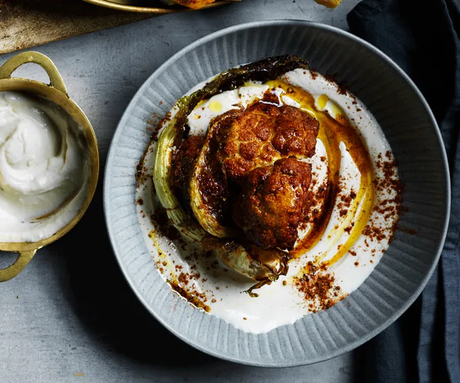Roasted cauliflower with creamy yogurt sauce and spices on a gray plate, next to a bowl of extra sauce.