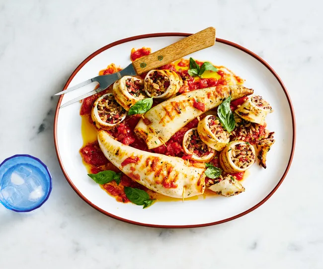 Grilled stuffed calamari with rice and herbs on a white plate with tomato sauce, garnished with basil leaves.