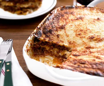 A white baking tray with lasagne, with a rectangle slice cut out. 