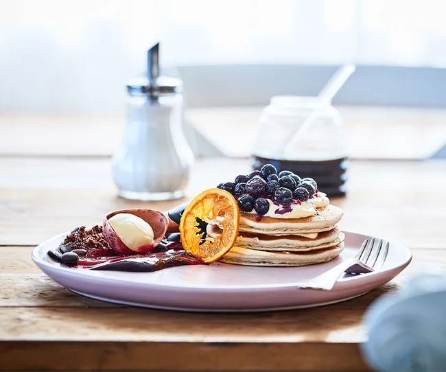 Pancakes with blueberries and orange slices on a plate, accompanied by cream, sauce, and a sugar dispenser in the background.