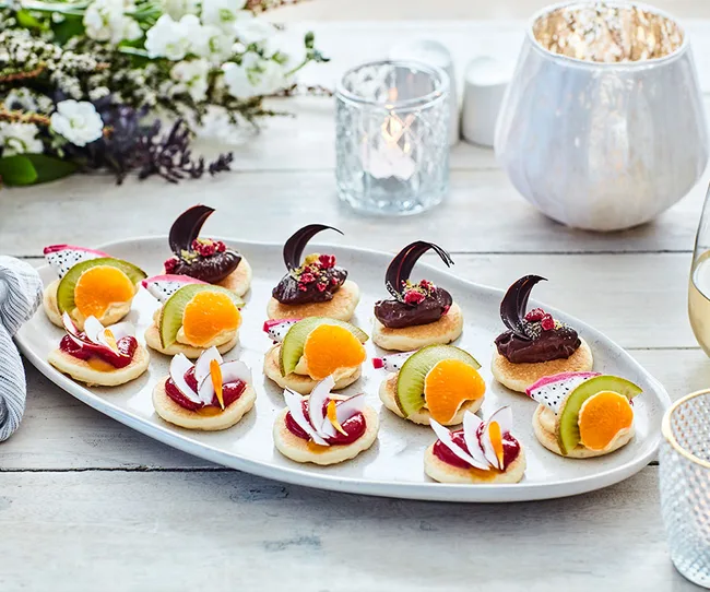 A platter of assorted gourmet canapés, including chocolate, fruit, and cream toppings, on a wooden table with candles.