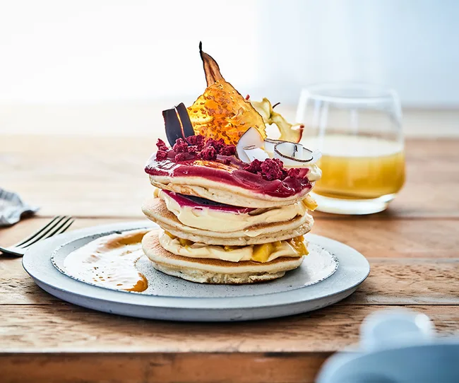 A stack of gourmet pancakes topped with fruit, chocolate, and sauce, served on a plate with a glass of juice in the background.