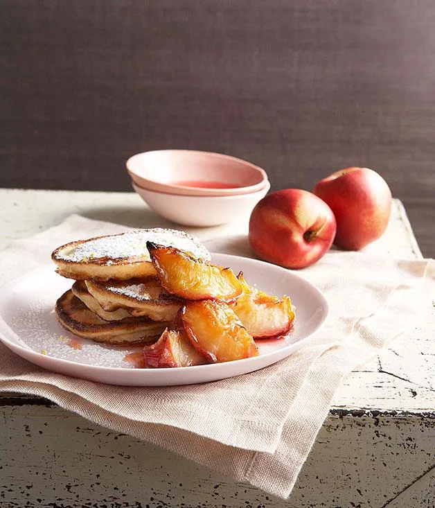 A plate of pancakes topped with sliced peaches on a rustic table, with whole peaches and a bowl in the background.