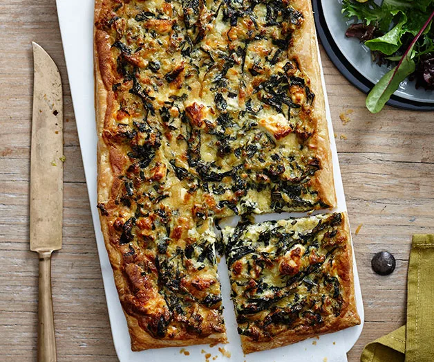 Rectangular kale and cheese tart on a white platter, one piece cut, next to a knife and salad.