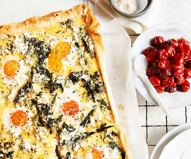 "Rectangular spinach and egg tart on a wire rack, served with a bowl of roasted cherry tomatoes."