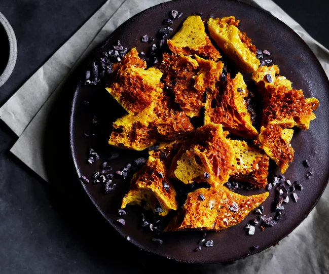 Honeycomb toffee pieces on a dark plate, sprinkled with cacao nibs, on a black and gray background.