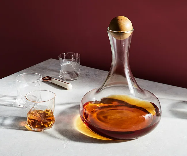 Decanter with amber liquid, wooden stopper, and glasses on a table with red wall background.