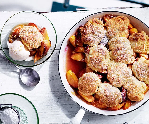 Peach cobbler in a baking dish with a serving in a bowl topped with ice cream on a white wooden table.