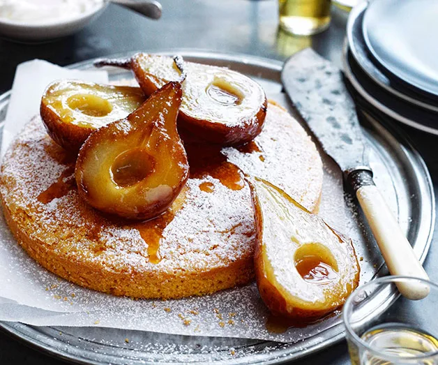 Caramelized pear halves on a round cake dusted with powdered sugar, served on a platter with a knife.
