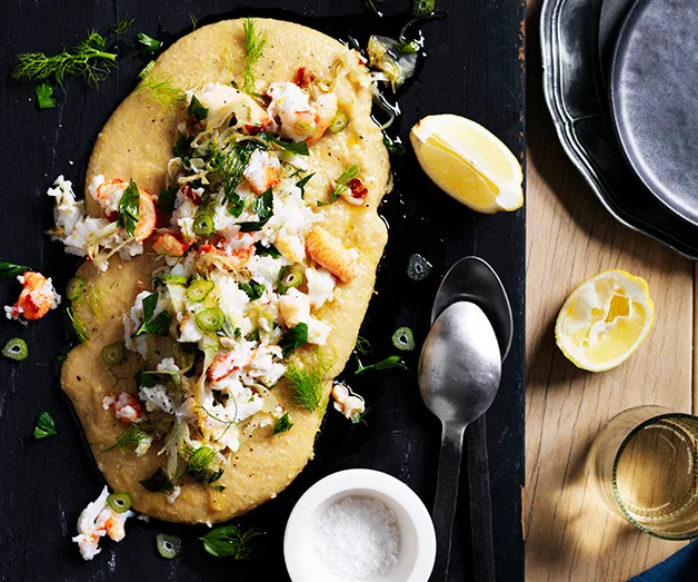 Polenta topped with crab, herbs, and green onions on a black board, lemon wedges, spoons, and salt on the side.