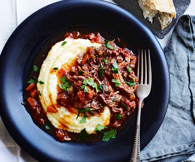 Braised beef ragu on creamy polenta, garnished with parsley, served in a black bowl with a fork and bread on the side.