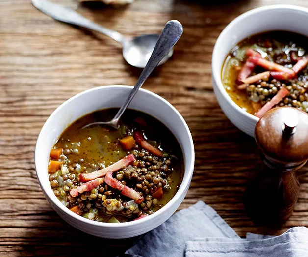 Two white bowls of lentil soup with bacon strips on a wooden table, accompanied by a spoon and pepper mill.