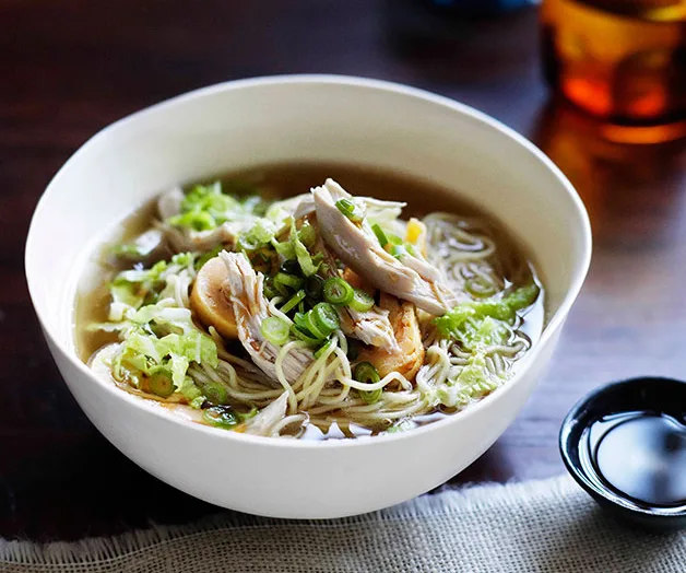 Bowl of chicken soba noodle soup topped with green onions and vegetables on a dark wooden table.