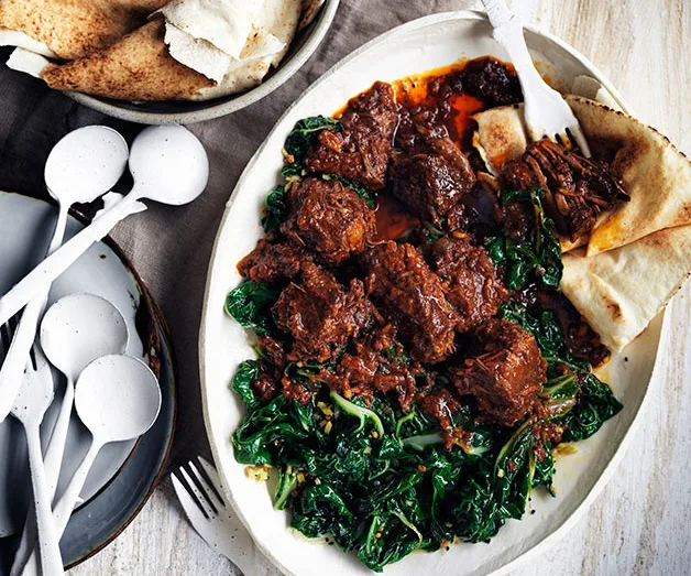 Plate of braised beef with greens, served with flatbread and white utensils on the side.