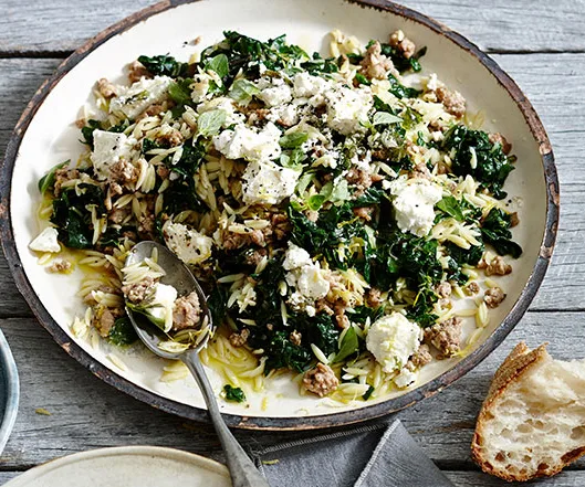Plate of orzo with sausage, kale, and ricotta, garnished with herbs, served with a slice of bread on a rustic wooden table.