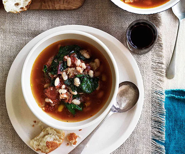 Bowl of vegetable soup with spinach, beans, and tomatoes on a table, served with bread and a small glass of red wine.