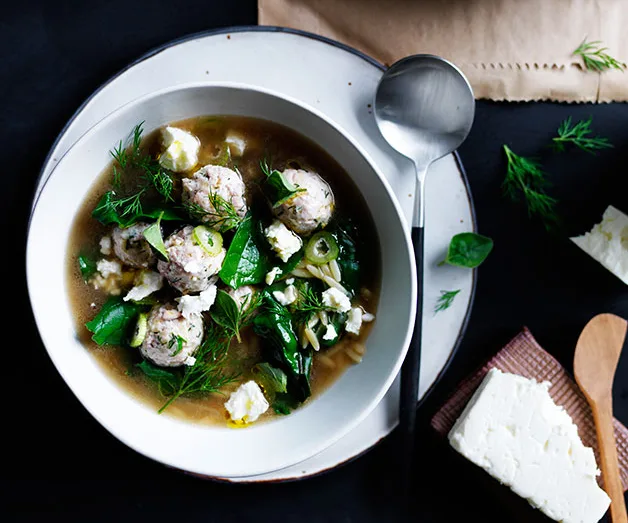 A bowl of soup with meatballs, spinach, dill, and feta cheese, garnished with herbs. A spoon and feta block beside the bowl.