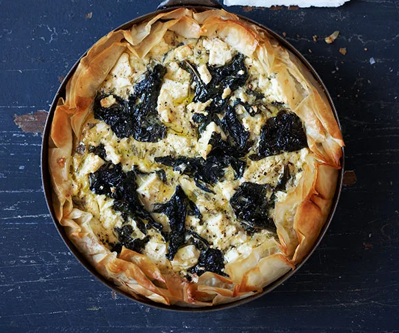 A golden brown kale and feta pie with a crispy phyllo crust, placed on a dark textured surface.