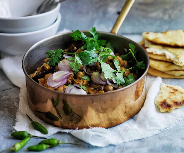 Copper pot of chickpea curry garnished with cilantro and onions, served with naan and green chilies on a white cloth.