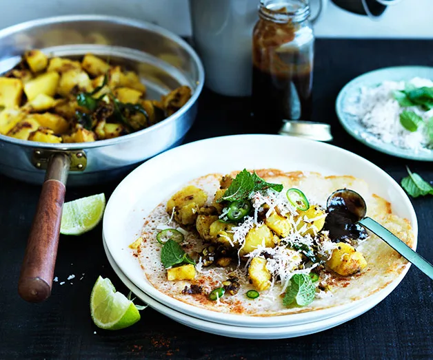 A dosa topped with spicy potatoes, garnished with mint and coconut, served with lime wedges and chutney.