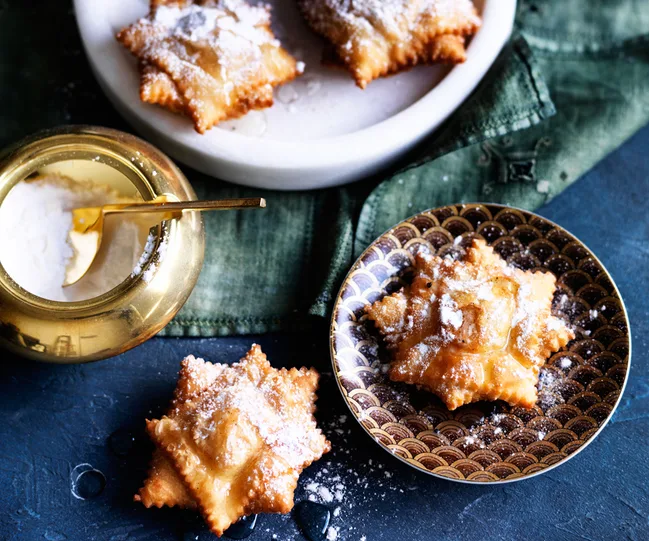 Star-shaped fried pastries dusted with powdered sugar on plates, next to a gold sugar bowl.