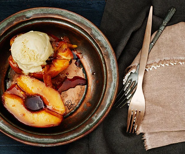 Roasted peach dessert with a scoop of vanilla ice cream in a rustic metal bowl, accompanied by a fork and knife on a napkin.