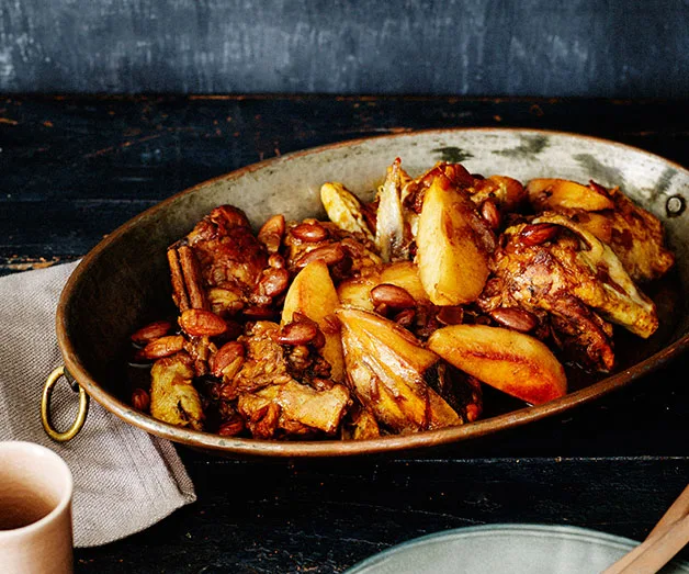 A roasted chicken dish with almonds and orange slices in a rustic metal dish on a dark wooden table.