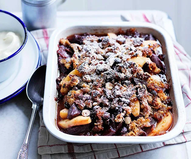 Rustic plum and apple crumble topped with oats in a baking dish, dusted with powdered sugar, with cream on the side.