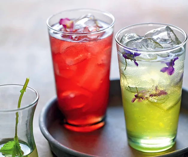 Two iced drinks: one red with flower garnish, another yellow-green on a gray tray.