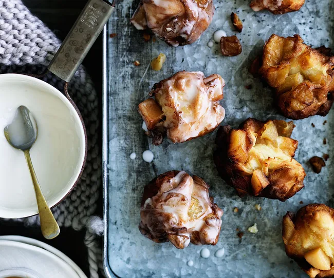 Apple fritters with icing on a baking tray, beside a bowl of cream with a spoon.